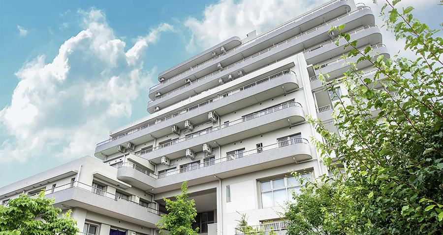 複数階の白色モダン建築の介護施設。青空の下、バルコニーと緑に囲まれた落ち着いた外観